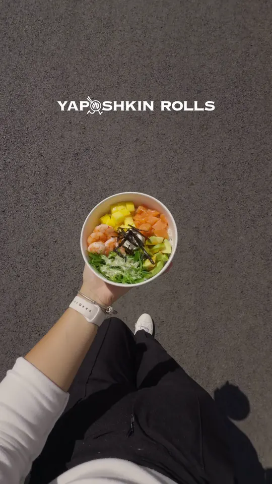 Seen from above on a sidewalk, a hand carries a poke bowl, then a tray of rolls.