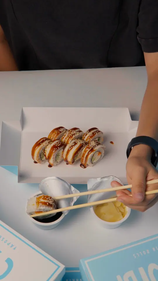 Hands open a light-blue Yaposhkin box of eel rolls, with cups of soy sauce and ginger beside it.